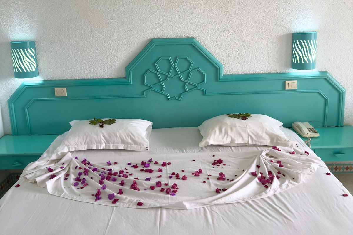 A bed with white linens and purple flower petals, a teal headboard, and a framed artwork above.