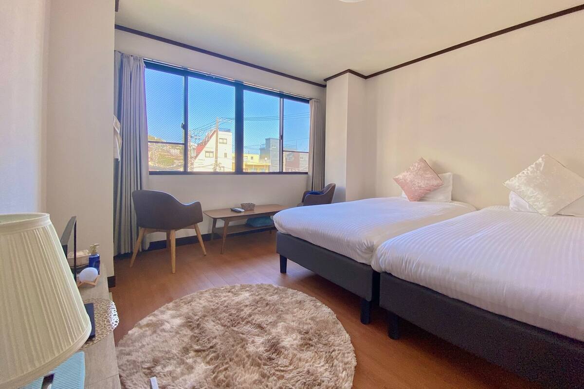 A hotel room with two beds, a desk, a chair, a round rug, and a large window offering a city view.