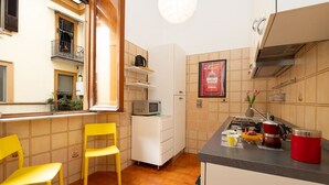 Eine kleine Küche mit Mikrowelle, Herd und einem roten Wasserkocher auf der Arbeitsfläche. Zwei gelbe Stühle und eine weiße Schrank stehen dort.