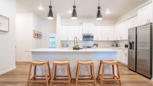 A modern kitchen with white cabinets, a stainless steel refrigerator, and a central island with stools. - Luxury Stay | Short Walk to AT&T Stadium + Parking (Arlington)