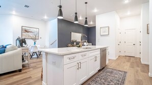 A modern kitchen with white cabinetry, a central island, pendant lighting, and a dining area with a white sofa and chairs. - Luxury Stay | Short Walk to AT&T Stadium + Parking (Arlington)
