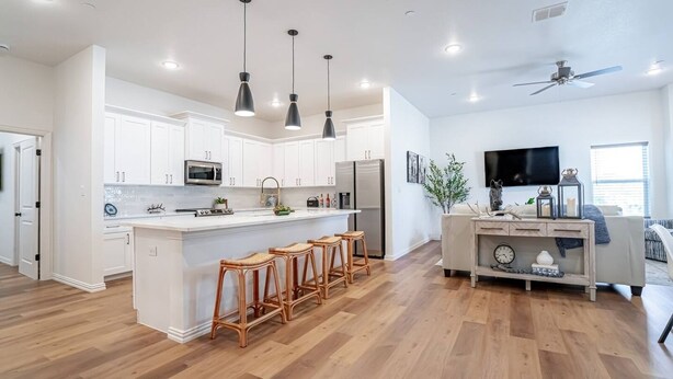 A modern kitchen with white cabinets, a central island with bar stools, and a dining area with a TV and decorative items. - Luxury Stay | Short Walk to AT&T Stadium + Parking (Arlington)