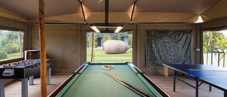 Une table de billard, une table de baby-foot et une table de ping-pong se trouvent dans une pièce aménagée sous une tente, avec vue sur un jardin.