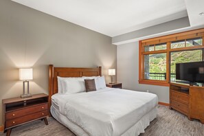 A bedroom with a large bed, wooden headboard, two bedside tables with lamps, a TV on a wooden cabinet, and a window with a view of greenery.