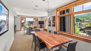 A modern dining area with a wooden table and chairs, large windows with a view of the outdoors, and a television in the background. - Gondola Grove | Capitol Peak Lodge #3310 | 2B/2B (Snowmass Village)