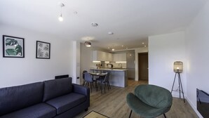 Ein modernes Wohnzimmer mit einem dunklen Sofa, einem grünen Sessel und einem Essbereich, im Hintergrund ist eine kleine Küche zu sehen.