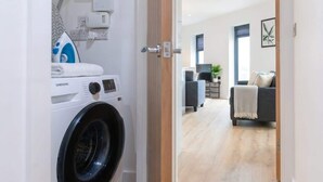 A laundry area with a Samsung washing machine, a drying machine, and a door leading to a living space with a sofa and a television.