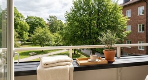 En balkon med udsigt over en park, en potteplante, en bakke med en kop og et sammenfoldet håndklæde.
