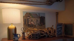 A wooden counter with a lamp, a ceramic pitcher, a spool of twine, a pile of rope, a painting of a house, and a chalkboard sign.