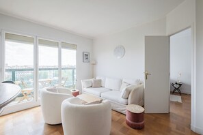 A modern living room with a white sofa, two white armchairs, a wooden coffee table, and a red ottoman. A large window offers a view of the city. - A Gleaming Vista, by Veeve (Paris)