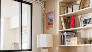 Ein ordentlich eingerichtetes Schlafzimmer mit einem Bett, einem Nachttisch, einer Lampe, einem Regal mit Büchern und Dekoration sowie einem Fenster mit Blick nach draußen.