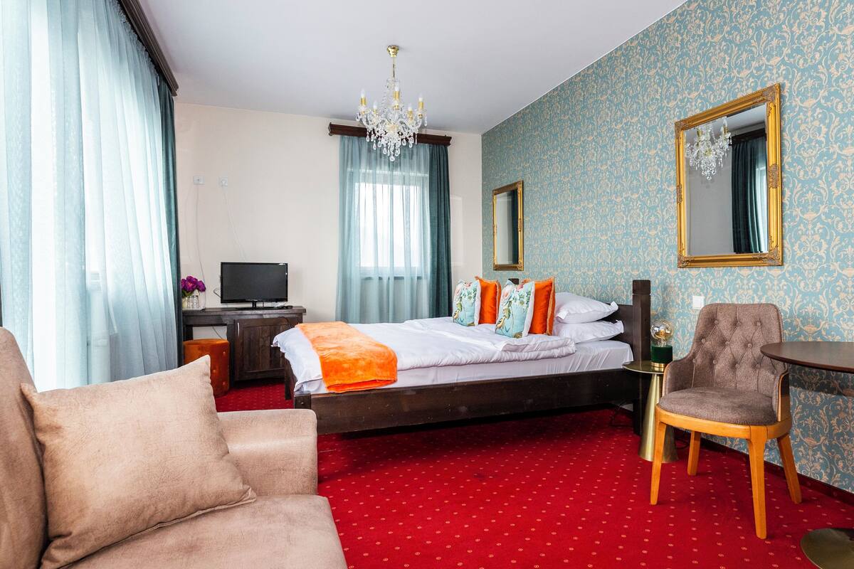 A hotel room with a bed, a TV, a desk, a chair, and a chandelier.