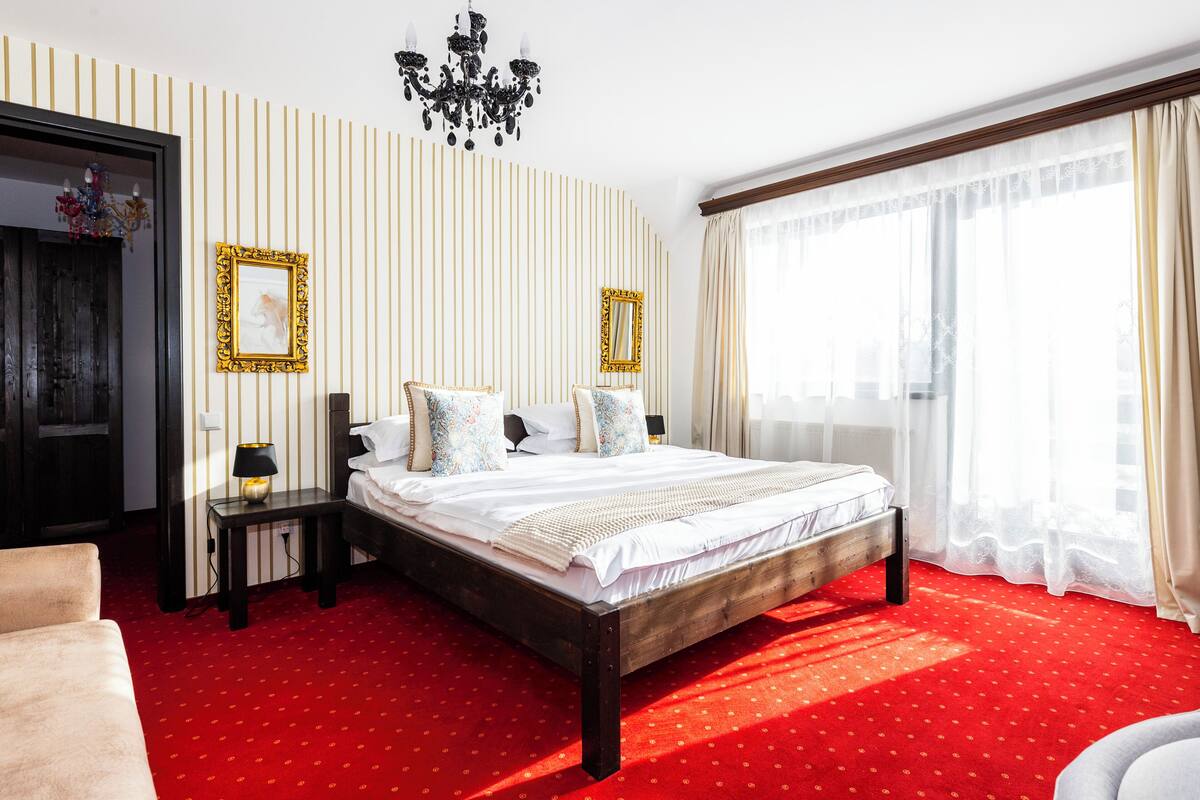 A hotel room with a large bed, a nightstand, a chair, a chandelier, and a window with curtains.