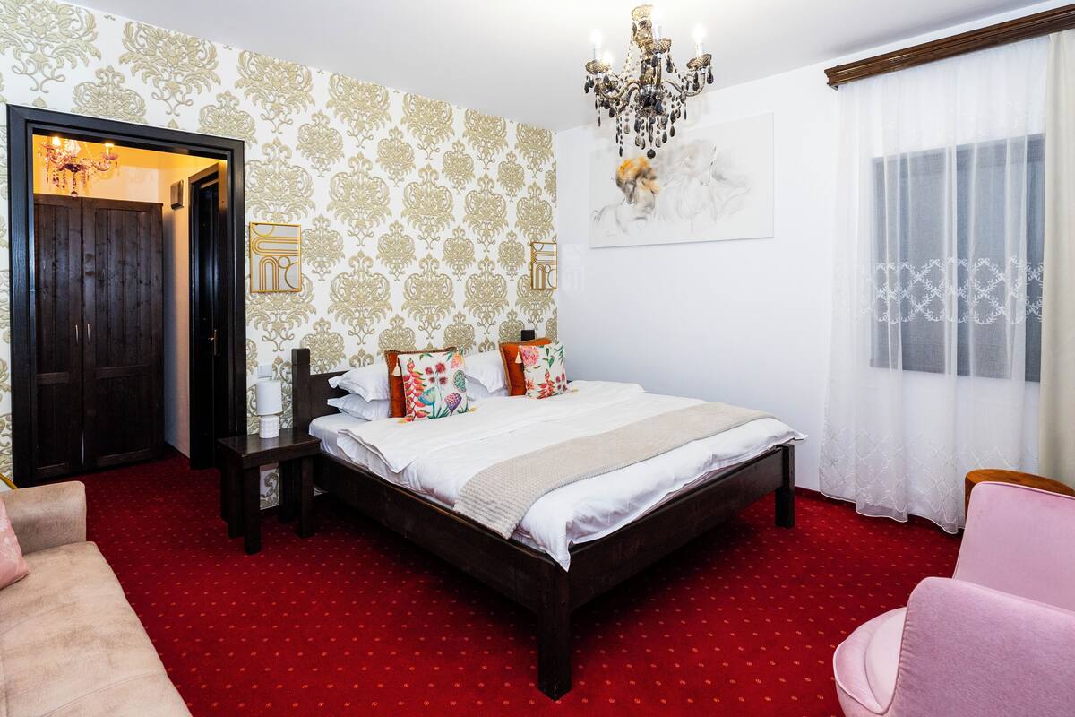 A bedroom with a large bed, a wooden wardrobe, a bedside table, a pink chair, and a red carpet.