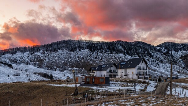 A two-story house with a balcony, surrounded by snow-covered hills and a sunset sky.