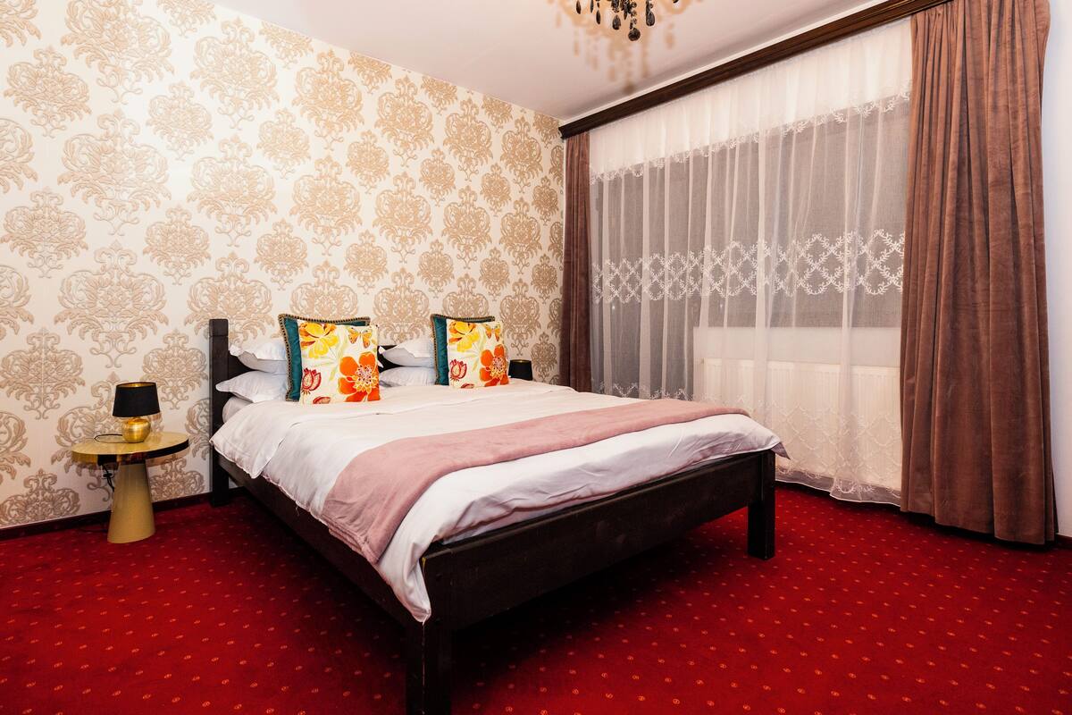A hotel room with a bed, a round side table with a lamp, a red carpet, and a large window with curtains.