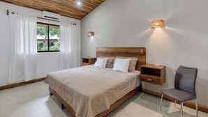 A bedroom with a wooden ceiling, a large bed with a headboard, two bedside tables, a chair, and a window with curtains. - Spacious 3-Bedroom Coastal Home with Shared Pool, Outdoor Living & Steps to Playa Potrero (Potrero)