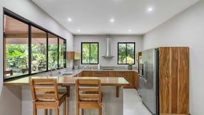 A modern kitchen with a central island, wooden chairs, a stainless steel refrigerator, and large windows offering a view of greenery. - Spacious 3-Bedroom Coastal Home with Shared Pool, Outdoor Living & Steps to Playa Potrero (Potrero)