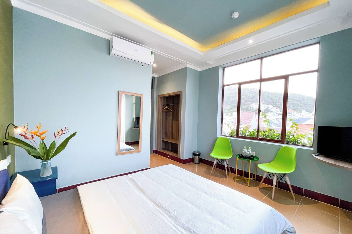 Una habitación de hotel con cama, espejo, ventana con vistas y dos sillones verdes.