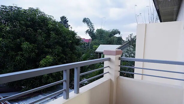 A balcony with a metal railing, a white wall, and a view of dense greenery and a building with a red roof. - Cozy, Safe, and Secure Two-Bedroom Apartment Unit (San Carlos City)
