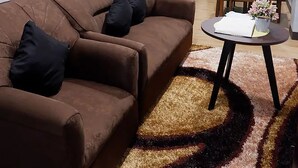A living room with a brown sofa, black cushions, and a round coffee table with a white cloth. A green dining area with a table and chairs is visible in the background. - Cozy, Safe, and Secure Two-Bedroom Apartment Unit (San Carlos City)