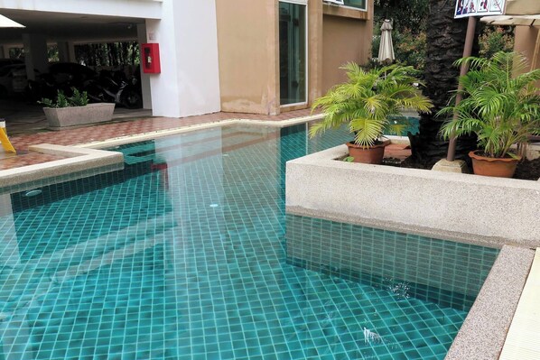 Une piscine dont le fond est recouvert de carreaux, entourée de plantes en pot et avec un bâtiment visible à l’arrière-plan.