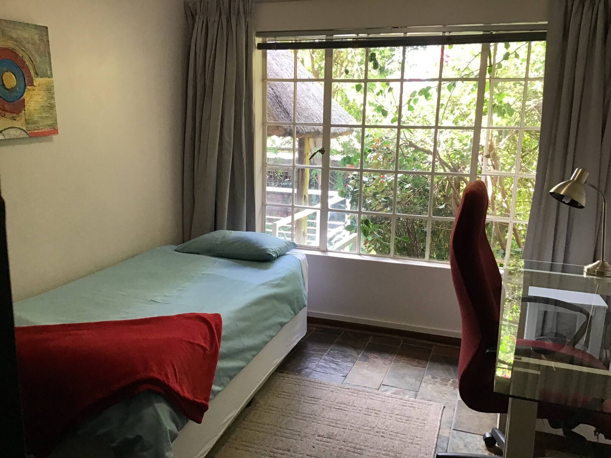 A room with a bed, a desk, a chair, a window with a view of greenery, and a painting on the wall.