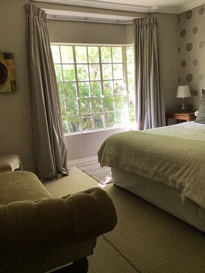 A bedroom with a large window, a bed, a bedside table with a lamp, and a chair.