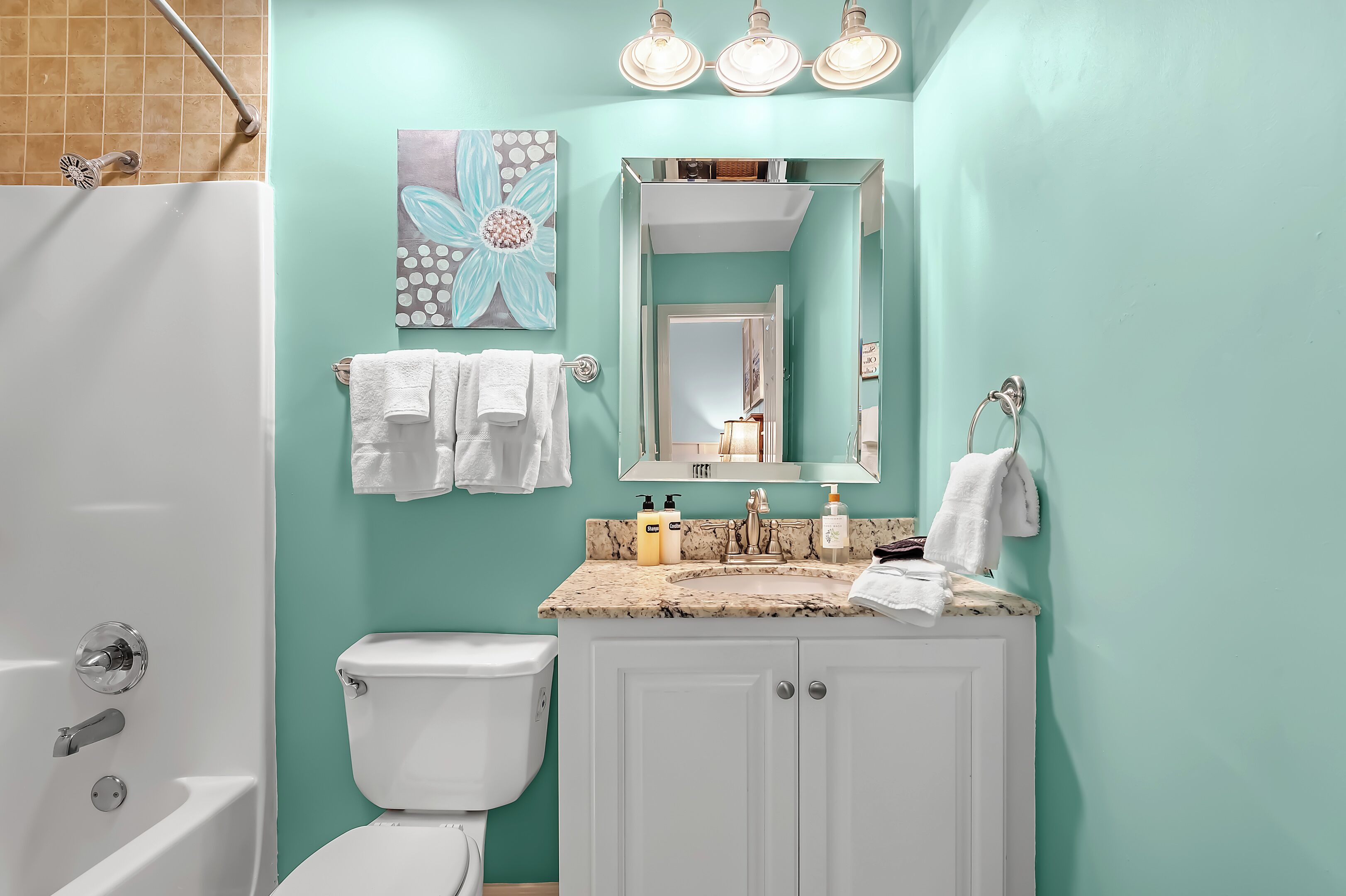 A bathroom with a white bathtub, a toilet, a sink with a marble countertop, and a mirror above the sink. There are three light fixtures above the mirror and a towel rack with towels.