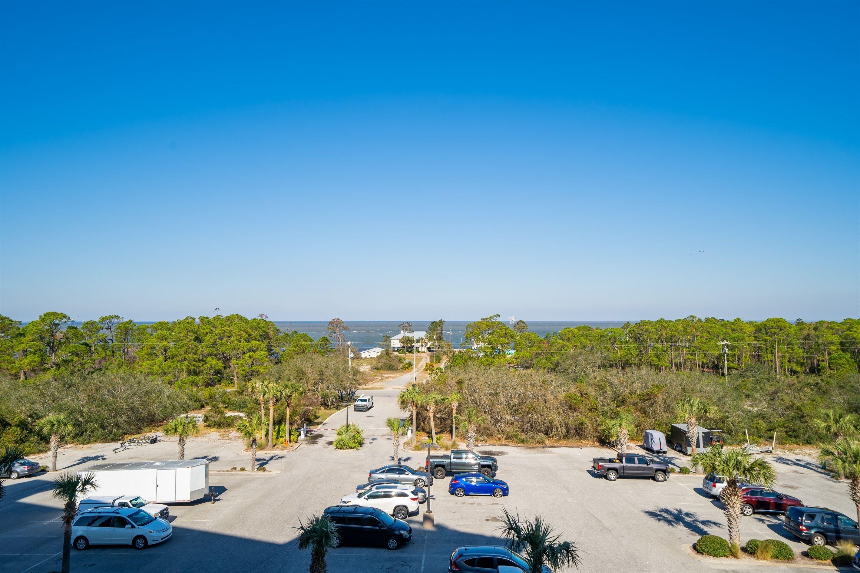 A parking lot with several cars and a trailer, surrounded by trees and a view of the ocean.
