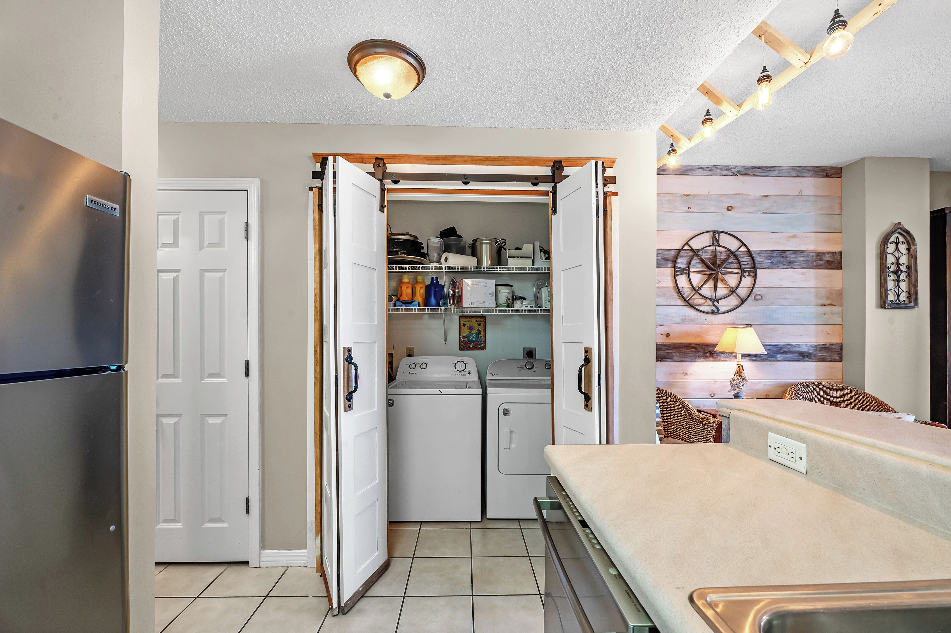 A compact kitchen with a refrigerator, an open door revealing a washing machine and dryer, a wooden wall with a decorative wheel, and a table with a lamp.