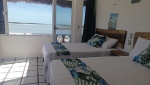 Een hotelkamer met twee bedden, uitzicht op zee, een rieten dak en een houten hoofdeinde.