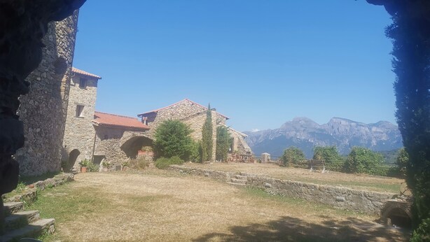 Un edificio de piedra con techo de tejas rojas, una entrada en arco y vistas a montañas al fondo.