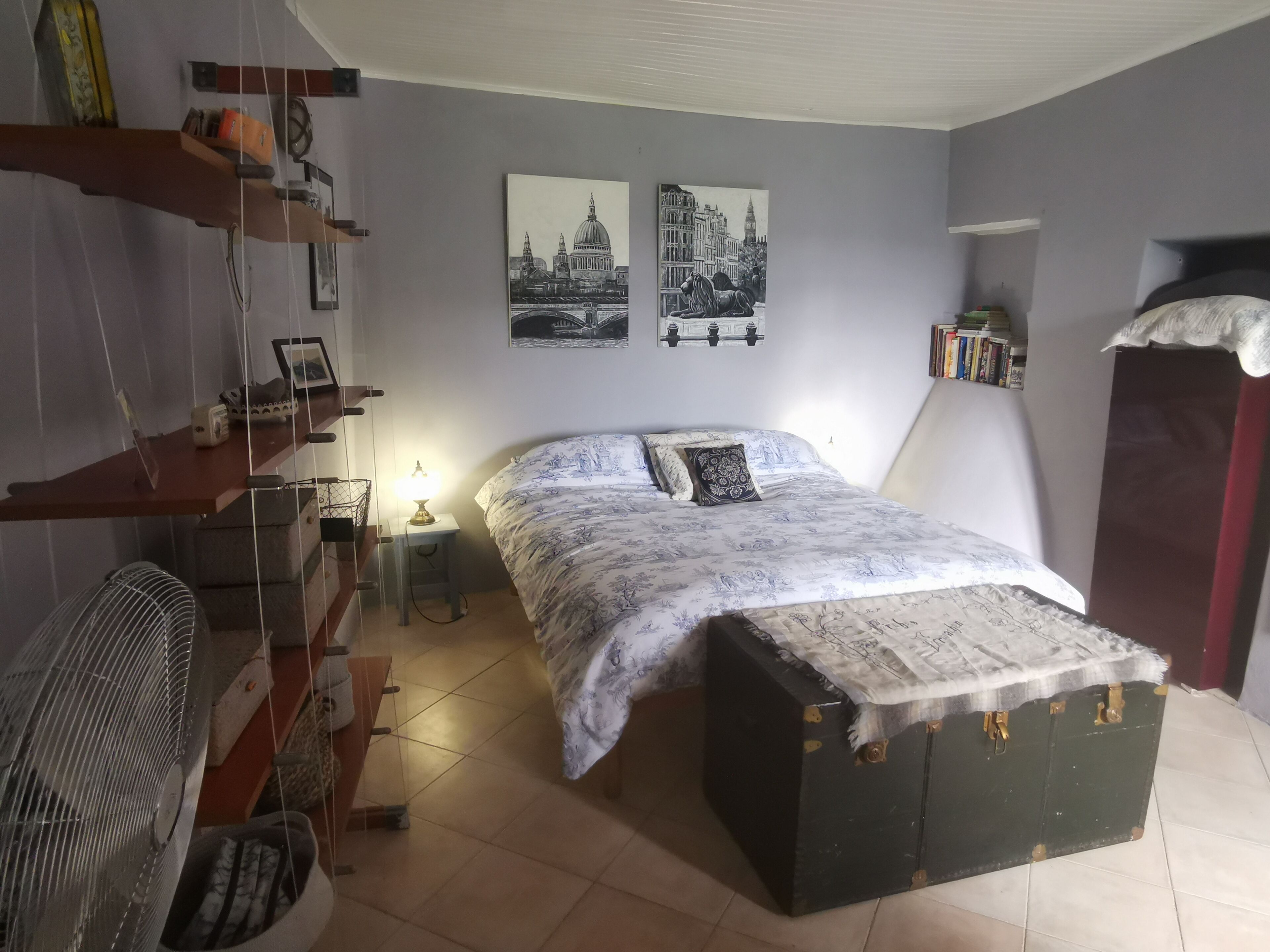 Un dormitorio con cama, escritorio, silla, ventilador y dos cuadros en la pared.