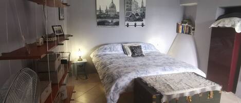 Un dormitorio con cama, escritorio, silla, ventilador y dos cuadros en la pared.