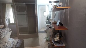 Habitación de hotel con cama, escritorio, silla, armario, estante con objetos y baño con inodoro y ducha.
