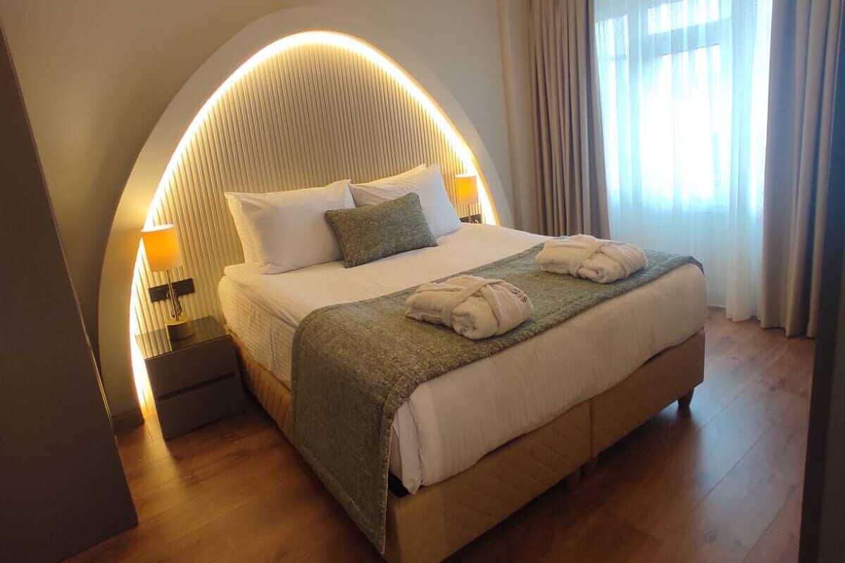 A modern hotel room with a curved ceiling light, a large bed with a green throw blanket, and a nightstand with a lamp.