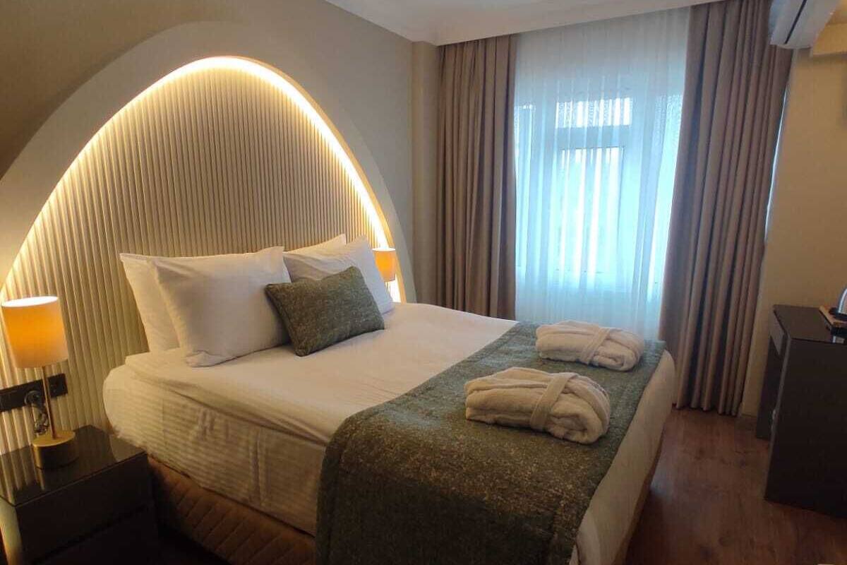 A modern hotel room with a curved headboard, a neatly made bed, a bedside table with a lamp, and a view of the outdoors through large windows.