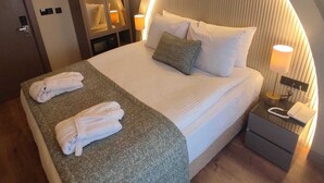 Une chambre d’hôtel moderne avec un grand lit, une tête de lit incurvée, un miroir, une table de chevet avec une lampe et un peignoir posé sur le lit.