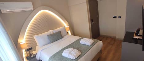 Une chambre d’hôtel moderne avec une tête de lit incurvée, un lit recouvert de draps blancs, un plaid vert et une table de chevet avec une lampe.