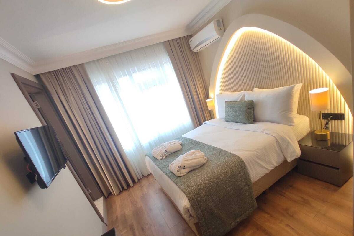 A hotel room with a bed, a TV, a bedside table with a lamp, and a window with curtains.