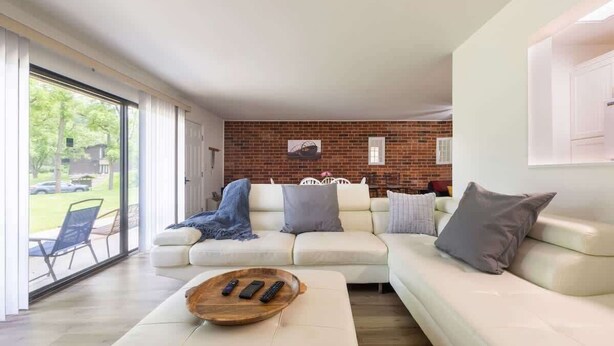 A modern living room with a white sofa, a wooden tray with remotes, a brick wall, and a glass door leading outside. - Charming 1BR Condo with Patio, Near The Lake Geneva (Lake Geneva)