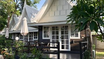 A traditional Thai house with a sloped roof, surrounded by lush greenery and a paved pathway.