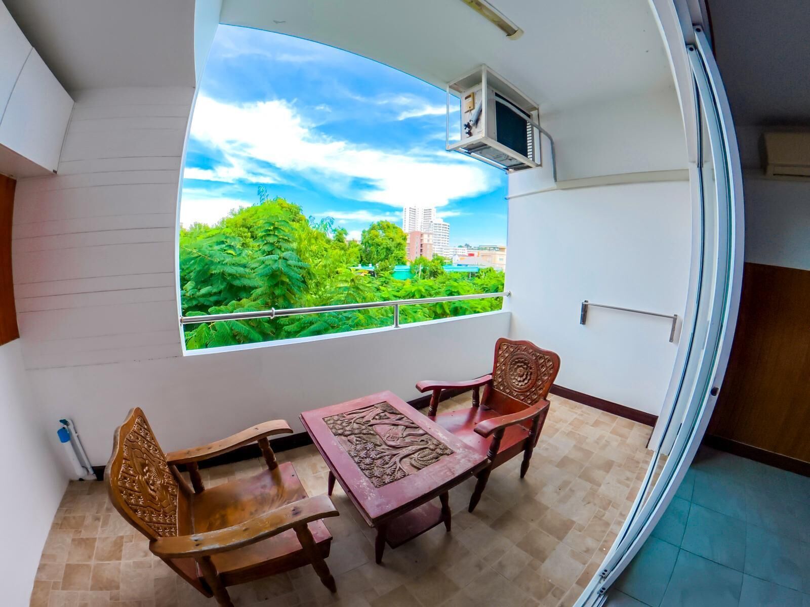 A balcony with a view of greenery and buildings, a table with chairs, and an air conditioning unit.