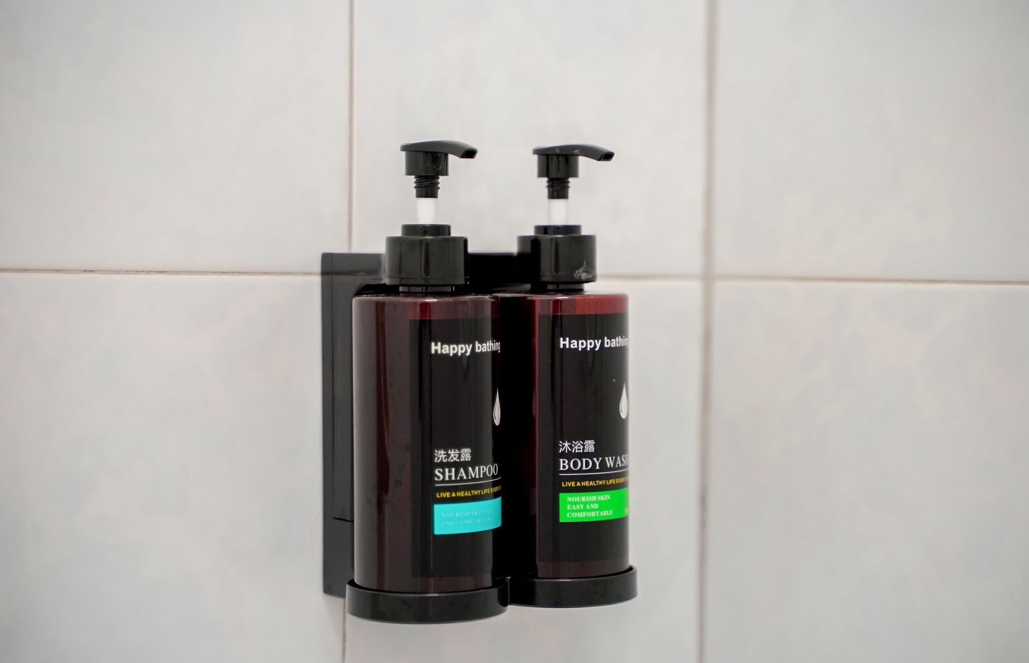 Two black bottles of Happy Bathing body wash and shampoo with pumps, mounted on a tiled wall.