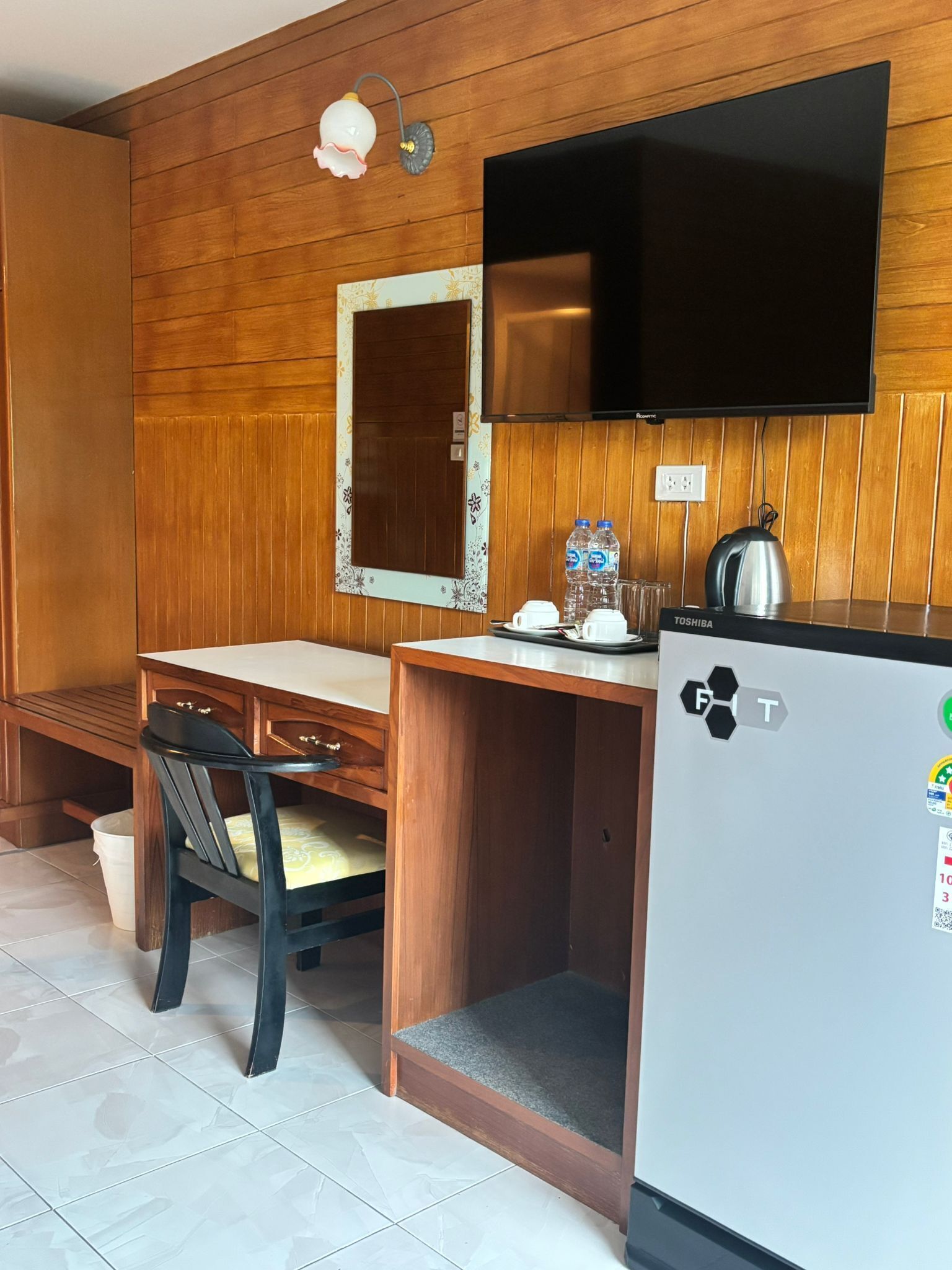 Une pièce de taille modeste avec un mur en bois, un téléviseur à écran plat fixé au-dessus d’un bureau en bois, un réfrigérateur et un banc.