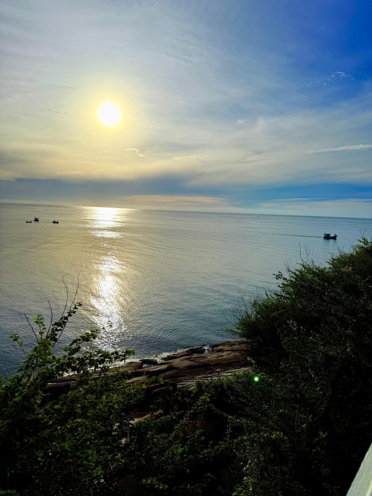A coastal view with the sun reflecting on the water, boats, and a rocky shoreline with vegetation.