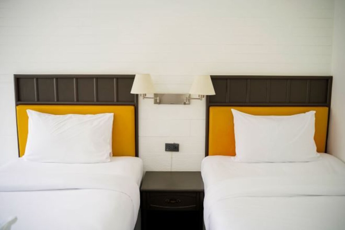 A hotel room with two beds, each with white bedding and a brown headboard. There are two bedside tables with lamps, and a wall-mounted light fixture above the beds.
