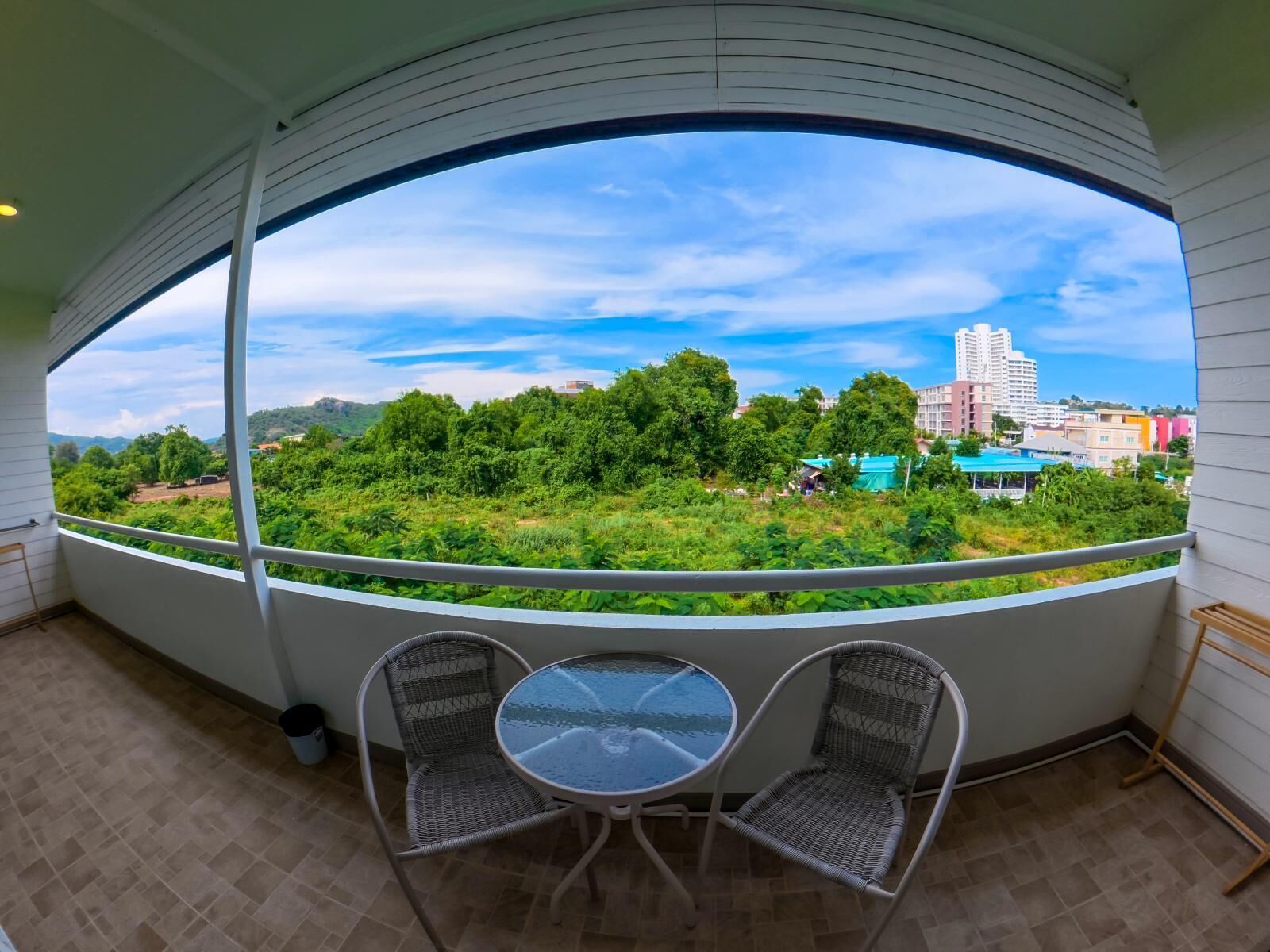 A balcony with a glass table and two wicker chairs, overlooking a green landscape and buildings.