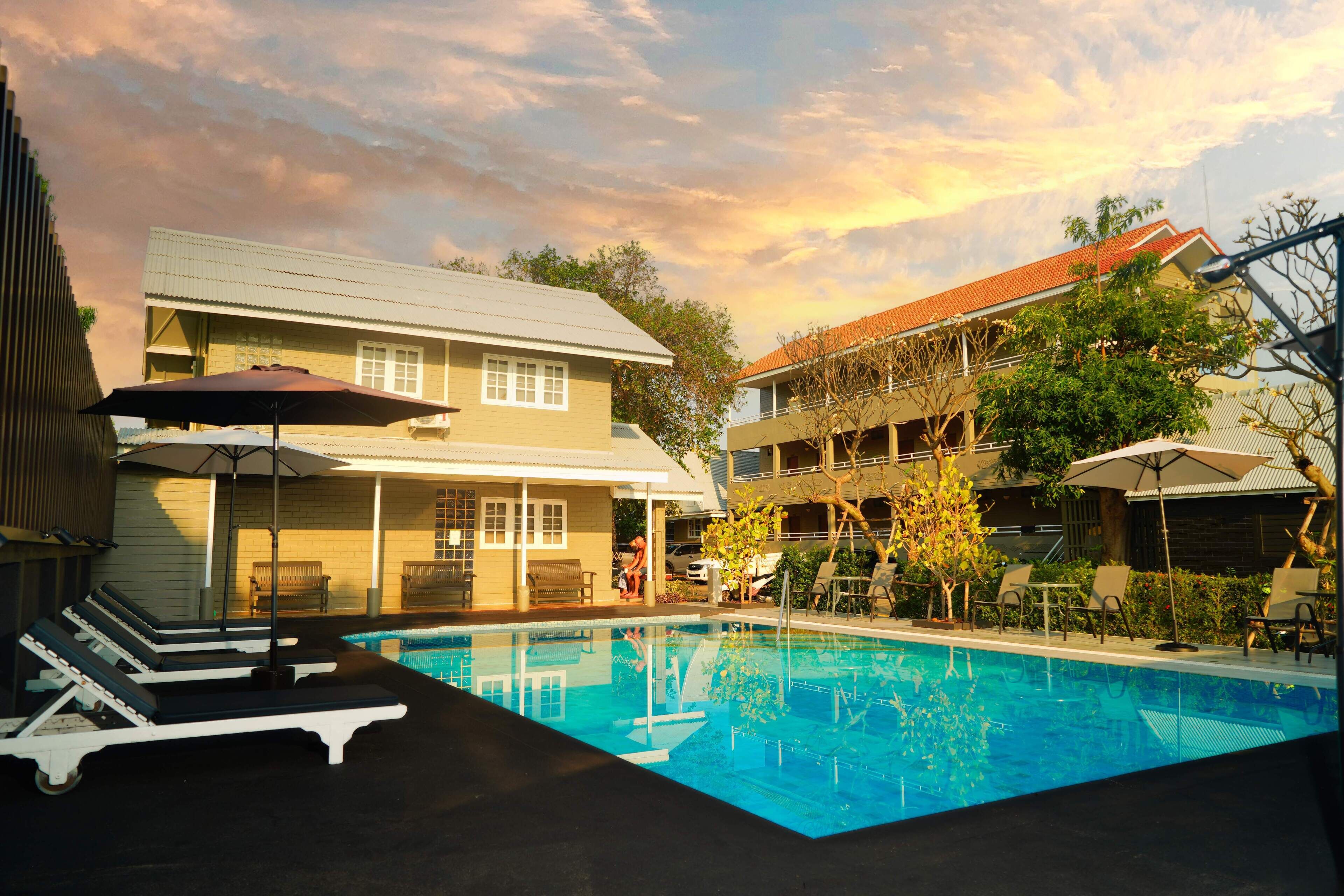 A swimming pool with lounge chairs and umbrellas, surrounded by buildings with balconies and trees.